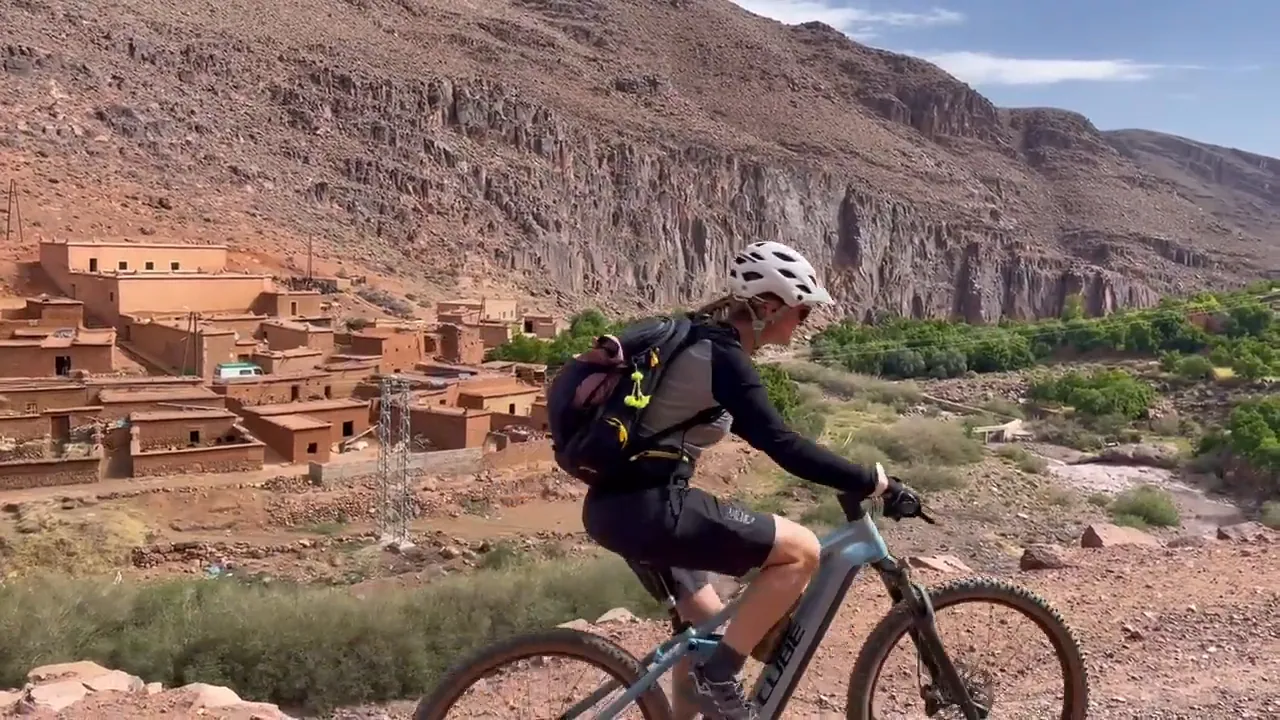 Riders pass a mud-brick village below a red cliff – cycling tour in Morocco